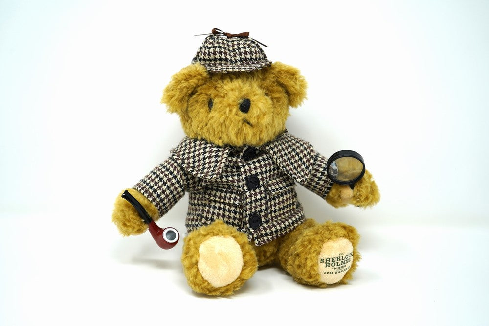 A small teddy bear dressed up as Sherlock Holmes, with a pipe in one paw, and a magnifying glass in the other.